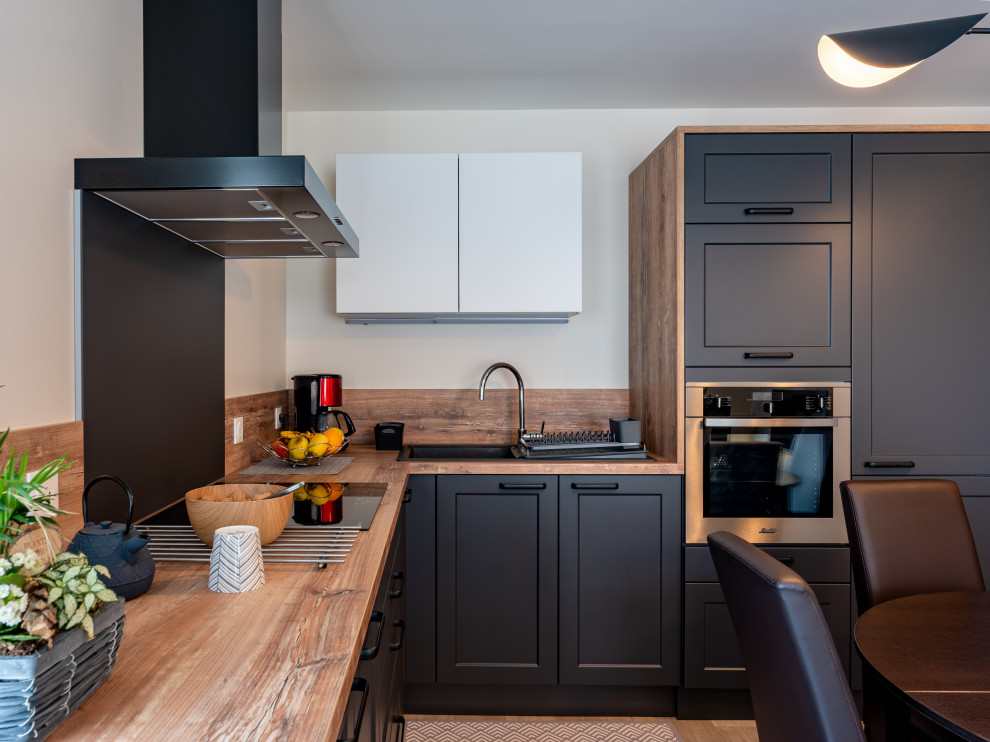 This is an example of a medium sized industrial l-shaped kitchen/diner in Other with an integrated sink, black cabinets, laminate countertops, beige splashback, wood splashback, stainless steel appliances, light hardwood flooring, no island, beige floors and beige worktops.