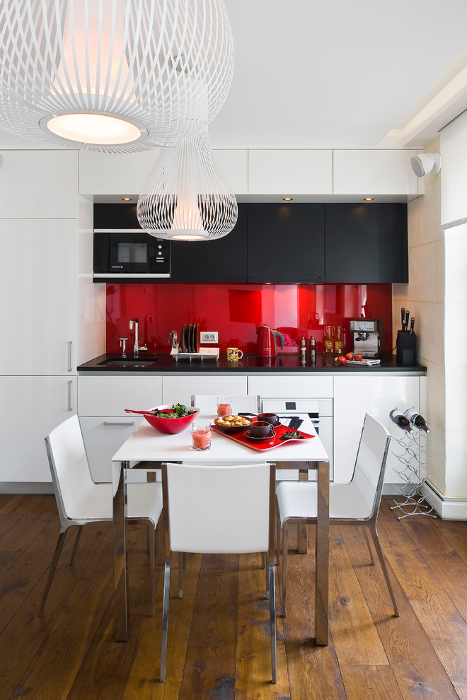 Cette image montre une cuisine américaine linéaire et encastrable design de taille moyenne avec des portes de placard blanches, une crédence rouge, une crédence en carreau de verre et un sol en bois brun.