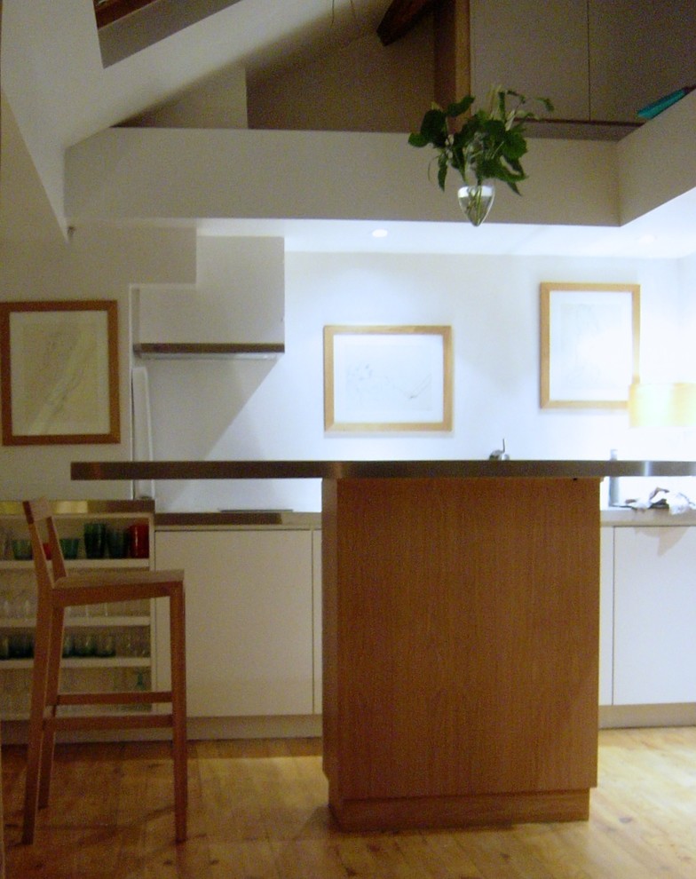 Bild på ett litet funkis linjärt kök och matrum, med en integrerad diskho, luckor med profilerade fronter, vita skåp, bänkskiva i rostfritt stål, vitt stänkskydd, rostfria vitvaror, ljust trägolv, en köksö och beiget golv