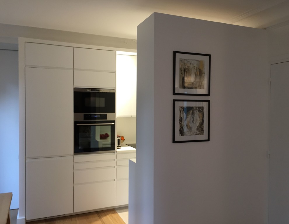 Cette image montre une petite cuisine ouverte parallèle design avec un évier intégré, un placard à porte affleurante, des portes de placard blanches, un plan de travail en surface solide, une crédence blanche, un électroménager noir, un sol en carrelage de céramique, îlot et un sol beige.