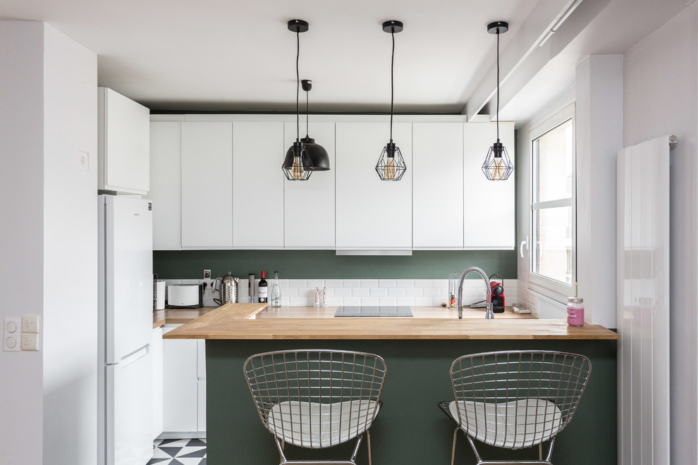 Bild på ett avskilt minimalistiskt l-kök, med släta luckor, vita skåp, träbänkskiva, vitt stänkskydd, stänkskydd i tunnelbanekakel, vita vitvaror, en halv köksö och svart golv