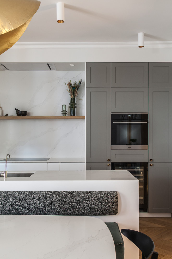Exemple d'une cuisine ouverte parallèle et encastrable scandinave avec un évier intégré, des portes de placard grises, un plan de travail en quartz, une crédence blanche, parquet clair, îlot et un plan de travail blanc.