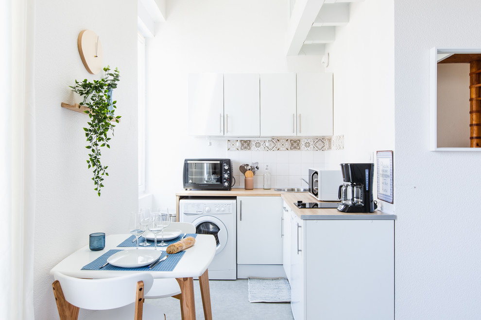 Пример оригинального дизайна: маленькая угловая кухня со стиральной машиной в морском стиле с обеденным столом, накладной мойкой, плоскими фасадами, белыми фасадами, разноцветным фартуком, серым полом, бетонным полом и окном для на участке и в саду