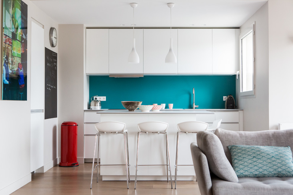 Cette image montre une cuisine ouverte linéaire design de taille moyenne avec un placard à porte plane, des portes de placard blanches, une crédence bleue, parquet clair et îlot.