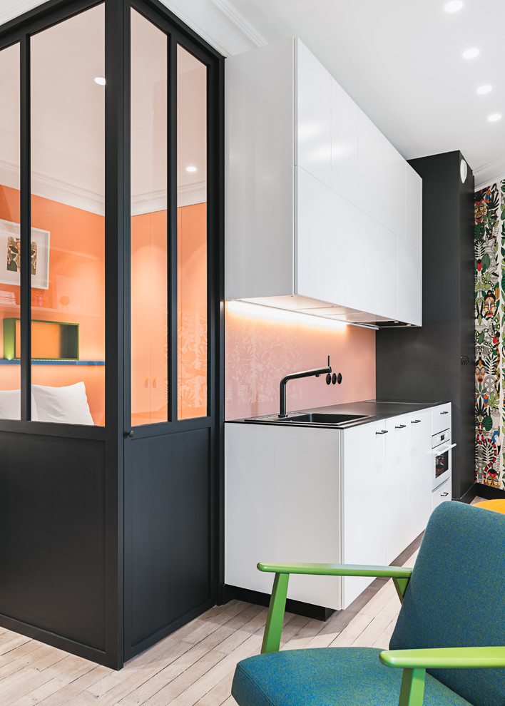Foto på ett funkis svart linjärt kök med öppen planlösning, med en nedsänkt diskho, släta luckor, vita skåp, rosa stänkskydd, glaspanel som stänkskydd, vita vitvaror, ljust trägolv och beiget golv