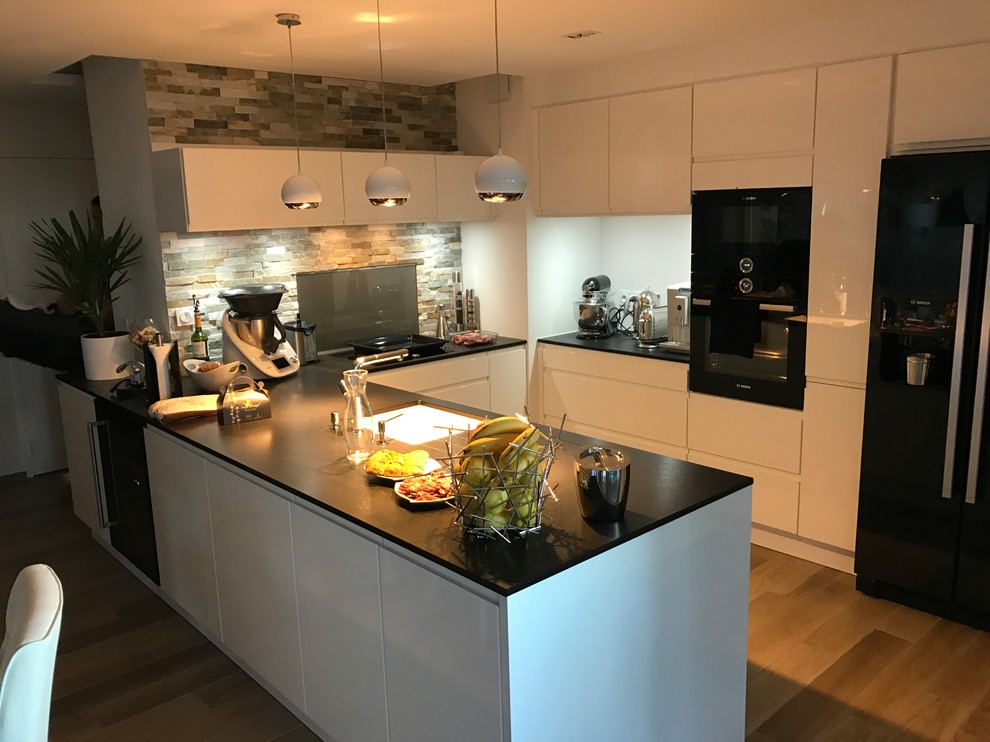Large contemporary u-shaped open plan kitchen in Lyon with a submerged sink, flat-panel cabinets, white cabinets, granite worktops, glass sheet splashback, black appliances, light hardwood flooring, an island, beige floors and black worktops.