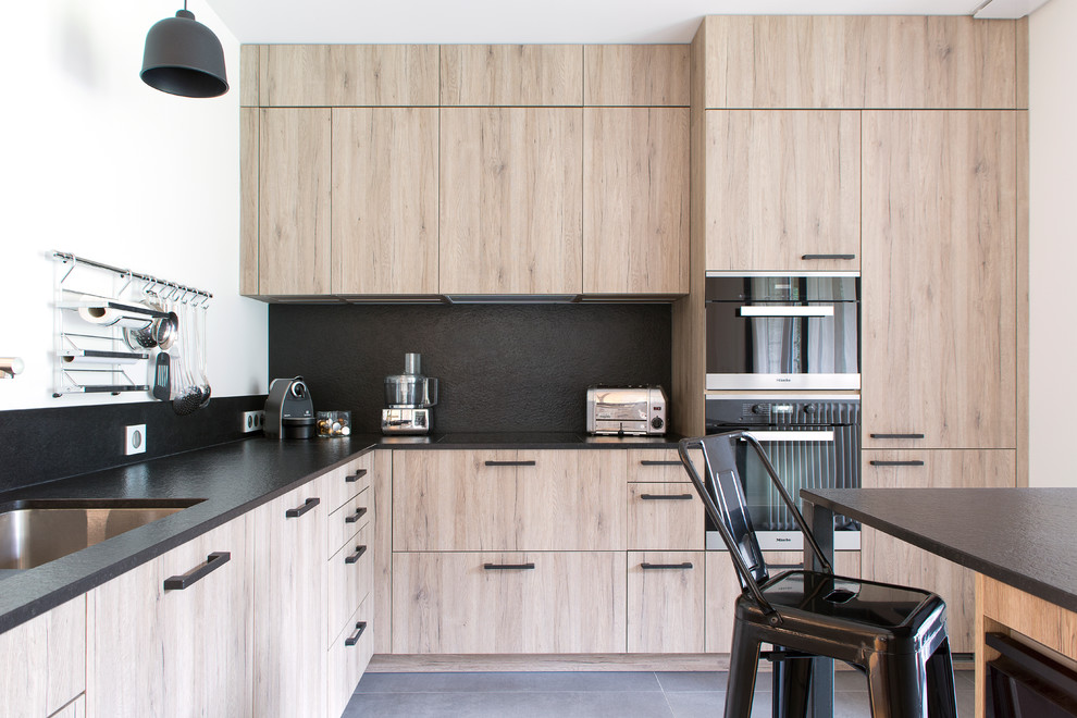 Exemple d'une cuisine scandinave en L et bois clair avec un plan de travail en granite, un évier encastré, un placard à porte plane, une crédence noire, un électroménager noir, îlot et un sol gris.