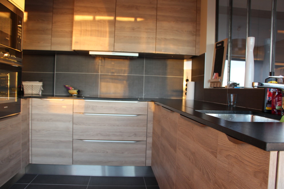 This is an example of a contemporary u-shaped open plan kitchen in Paris with a single-bowl sink, flat-panel cabinets, light wood cabinets, granite worktops, grey splashback, ceramic splashback, black appliances and ceramic flooring.