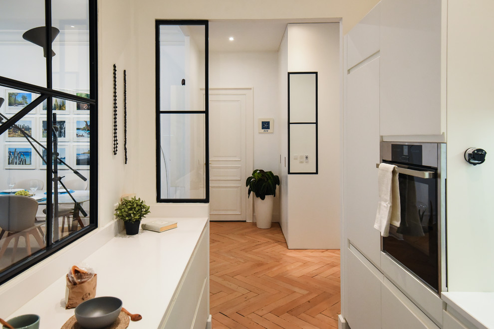 Moderne Küche mit Kassettenfronten, weißen Schränken, Küchenrückwand in Weiß, Küchengeräten aus Edelstahl, braunem Holzboden, Kücheninsel und beigem Boden in Lyon