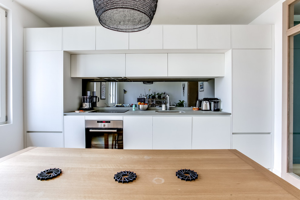 Inspiration för små minimalistiska linjära kök med öppen planlösning, med en undermonterad diskho, släta luckor, vita skåp, bänkskiva i kvartsit, grått stänkskydd, spegel som stänkskydd, integrerade vitvaror och ljust trägolv