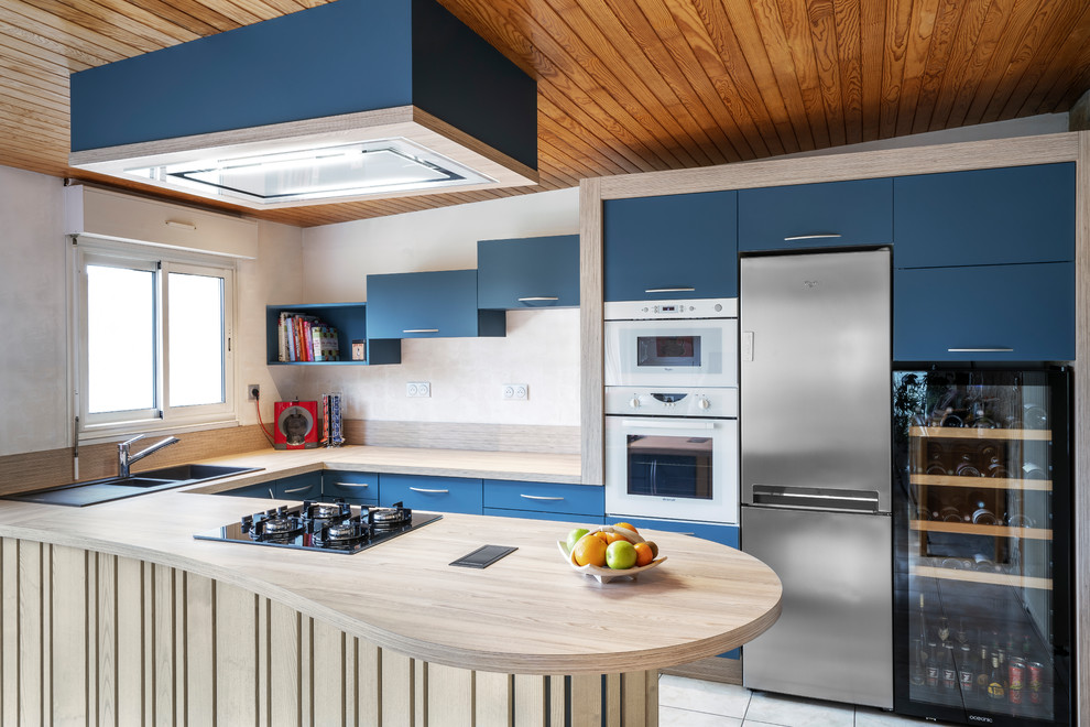 На фото: п-образная кухня в современном стиле с накладной мойкой, плоскими фасадами, синими фасадами, деревянной столешницей, полуостровом, серым полом и бежевой столешницей