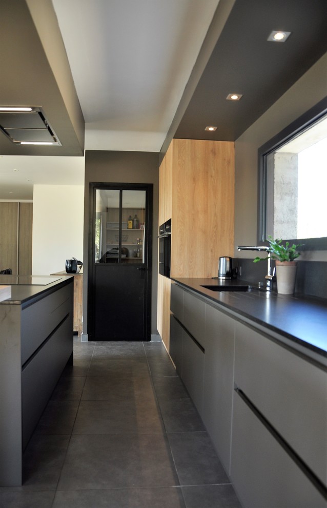 Exemple d'une cuisine parallèle tendance avec un plan de travail en granite, une crédence noire, un électroménager noir, un sol en carrelage de céramique, îlot, un sol gris et plan de travail noir.