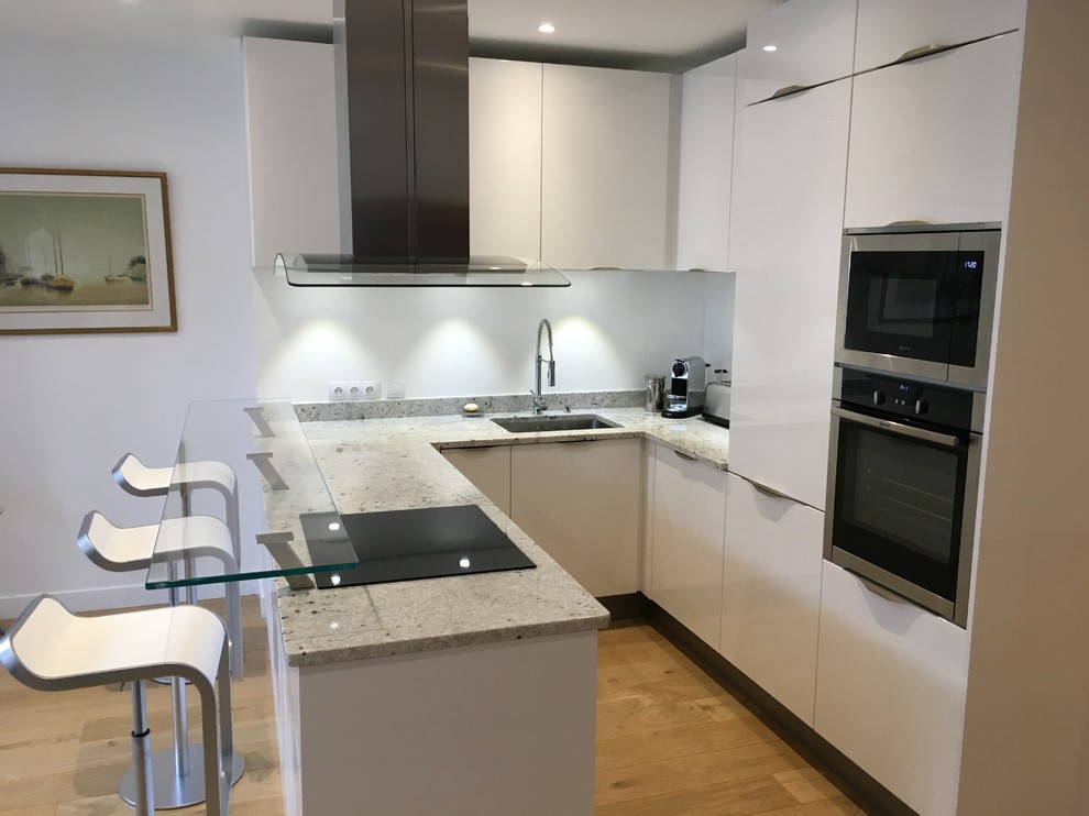 This is an example of a medium sized contemporary u-shaped kitchen/diner in Paris with an integrated sink, white cabinets, granite worktops, integrated appliances, light hardwood flooring, a breakfast bar, beaded cabinets, white splashback and beige floors.