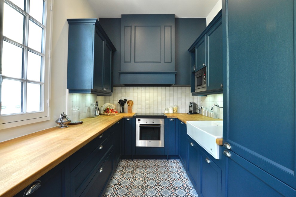 Klassisk inredning av ett avskilt, litet u-kök, med en integrerad diskho, skåp i shakerstil, blå skåp, träbänkskiva, vitt stänkskydd, stänkskydd i keramik, rostfria vitvaror och cementgolv