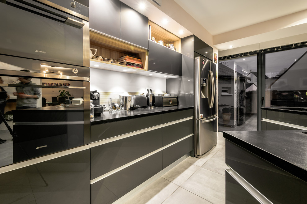 Large contemporary l-shaped open plan kitchen in Angers with grey cabinets, granite worktops, brown splashback, black appliances and an island.