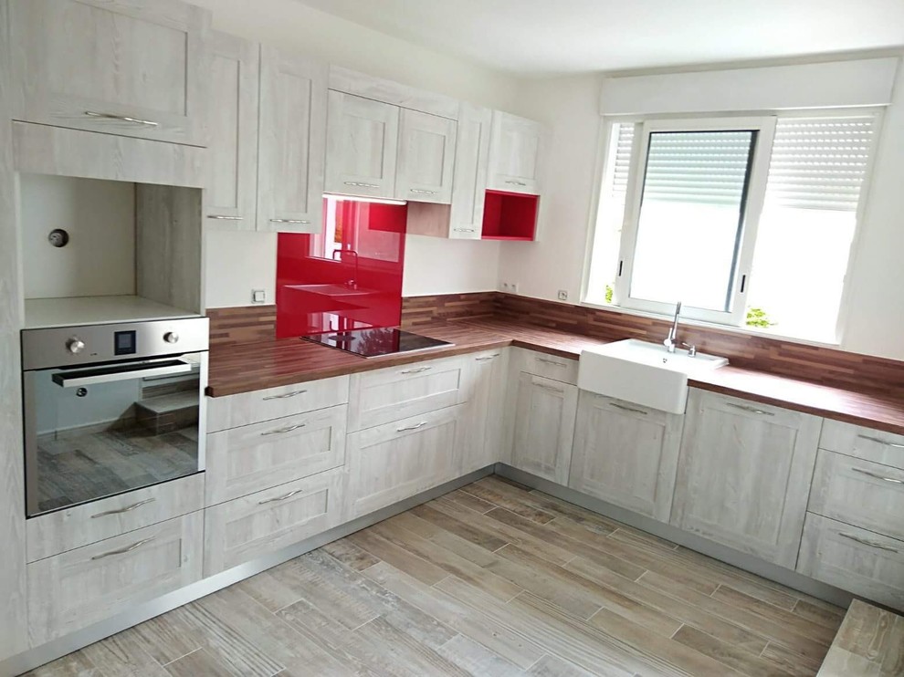 Photo of a medium sized retro l-shaped enclosed kitchen in Other with a belfast sink, beaded cabinets, distressed cabinets, laminate countertops, red splashback, mirror splashback, stainless steel appliances, laminate floors, no island, brown floors and brown worktops.