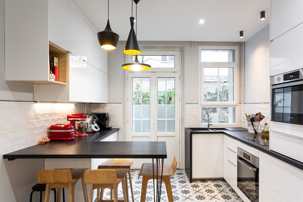 This is an example of a medium sized contemporary galley enclosed kitchen in Paris with a double-bowl sink, beaded cabinets, white cabinets, laminate countertops, white splashback, terracotta splashback, stainless steel appliances, cement flooring, grey floors, black worktops and a breakfast bar.