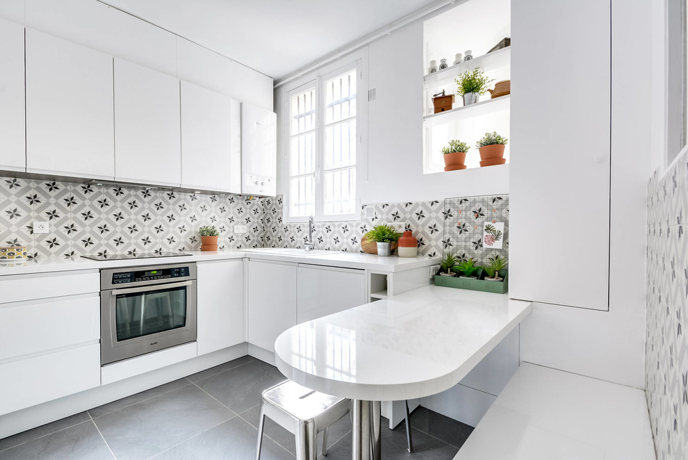 This is an example of a medium sized contemporary l-shaped kitchen/diner in Montpellier with a built-in sink, flat-panel cabinets, white cabinets, multi-coloured splashback, cement tile splashback, stainless steel appliances, grey floors, laminate countertops, ceramic flooring, an island and white worktops.