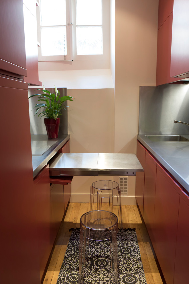 This is an example of a small contemporary galley enclosed kitchen in Paris with an integrated sink, beaded cabinets, red cabinets, stainless steel worktops, grey splashback, integrated appliances, light hardwood flooring, brown floors and grey worktops.
