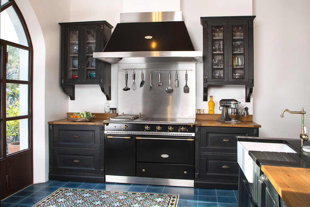 Klassische Küche mit Landhausspüle, profilierten Schrankfronten, schwarzen Schränken, Arbeitsplatte aus Holz, Küchenrückwand in Metallic, schwarzen Elektrogeräten, blauem Boden und brauner Arbeitsplatte in Marseille