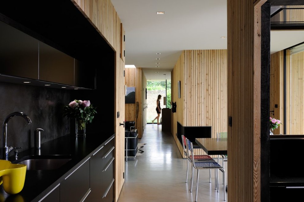 Источник вдохновения для домашнего уюта: кухня-гостиная среднего размера в современном стиле с одинарной мойкой, черными фасадами, гранитной столешницей, черным фартуком, техникой под мебельный фасад и бетонным полом