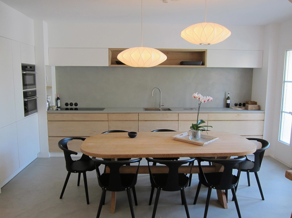 На фото: угловая кухня среднего размера в современном стиле с обеденным столом, врезной мойкой, светлыми деревянными фасадами, серым фартуком, техникой из нержавеющей стали и бетонным полом с