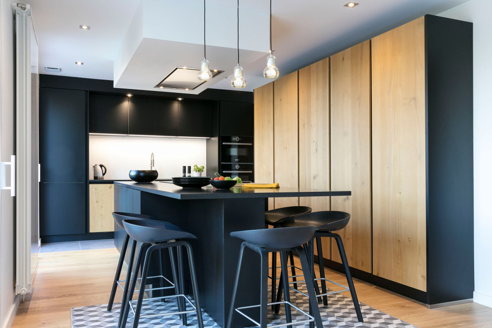 Cette image montre une cuisine design en L et bois clair avec un placard à porte plane, un électroménager noir, un sol en bois brun, îlot, un sol marron et plan de travail noir.