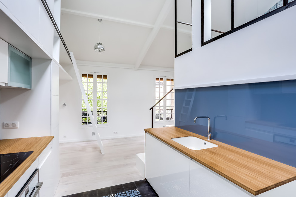 Small contemporary galley open plan kitchen in Paris with a submerged sink, beaded cabinets, white cabinets, wood worktops, blue splashback, glass sheet splashback, integrated appliances, cement flooring, no island and blue floors.