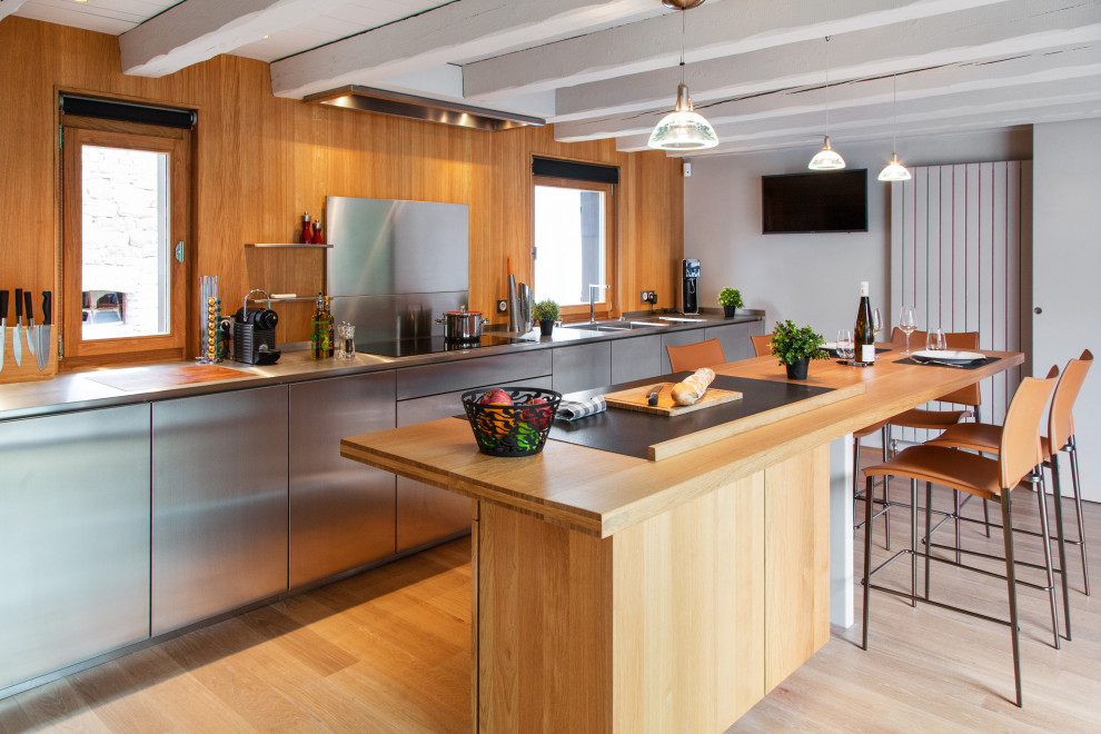 Cette photo montre une cuisine parallèle industrielle en inox avec un placard à porte plane, une crédence marron, une crédence en bois, parquet clair, îlot, un sol beige, un plan de travail gris et poutres apparentes.