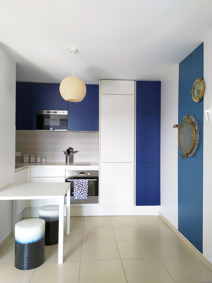 Cette image montre une cuisine marine en U avec un placard à porte plane, des portes de placard bleues, une crédence beige, un électroménager en acier inoxydable, une péninsule, un sol beige et un plan de travail blanc.