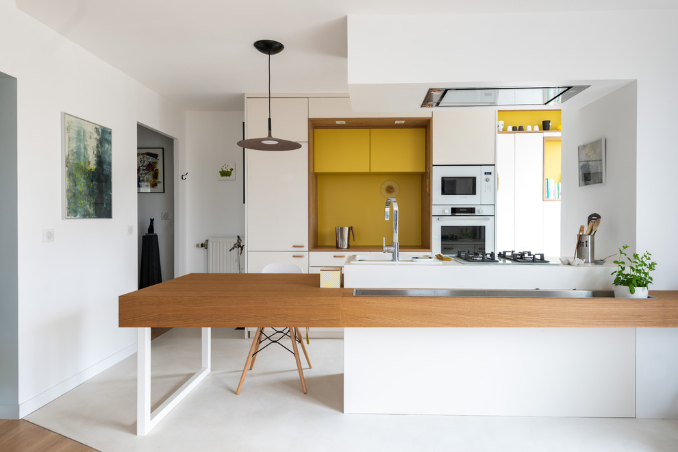 Foto på ett mellanstort funkis vit kök, med vita skåp, träbänkskiva, gult stänkskydd, vita vitvaror, betonggolv, en halv köksö, vitt golv och släta luckor