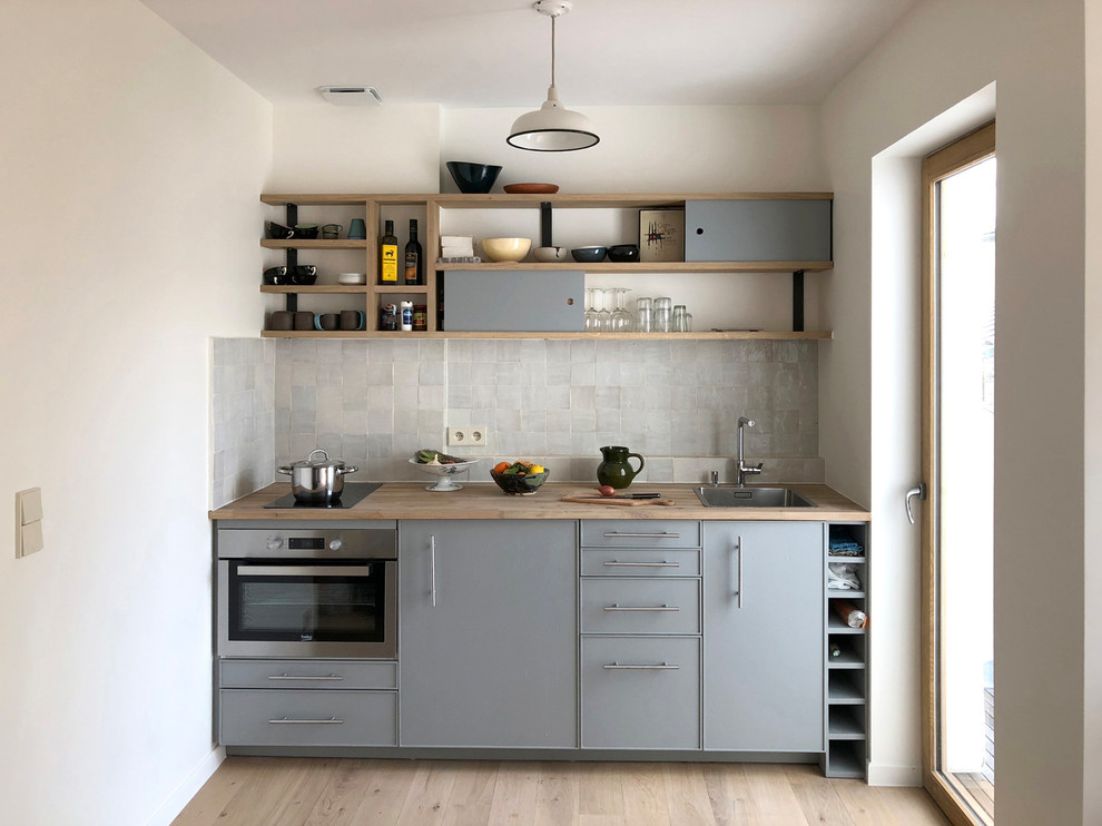 Exempel på ett litet minimalistiskt linjärt kök, med en enkel diskho, släta luckor, grå skåp, träbänkskiva, grått stänkskydd, rostfria vitvaror och ljust trägolv