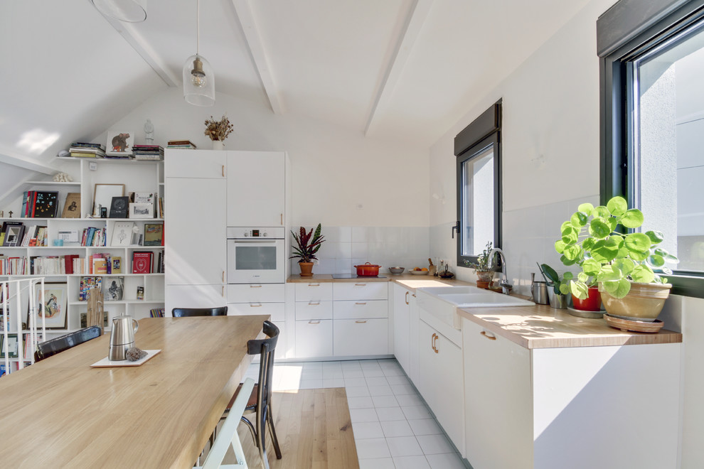 Contemporary l-shaped kitchen/diner in Paris with a belfast sink, flat-panel cabinets, white cabinets, wood worktops, white splashback, glass tiled splashback, white appliances, white floors and beige worktops.