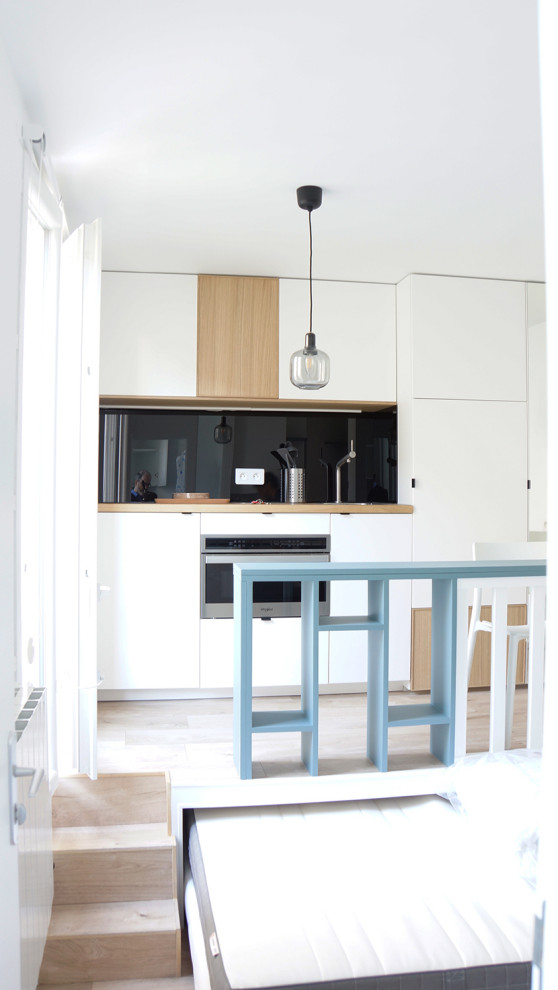 Design ideas for a small contemporary galley open plan kitchen in Paris with a single-bowl sink, beaded cabinets, white cabinets, wood worktops, black splashback, glass tiled splashback, stainless steel appliances, laminate floors, an island, brown floors and brown worktops.