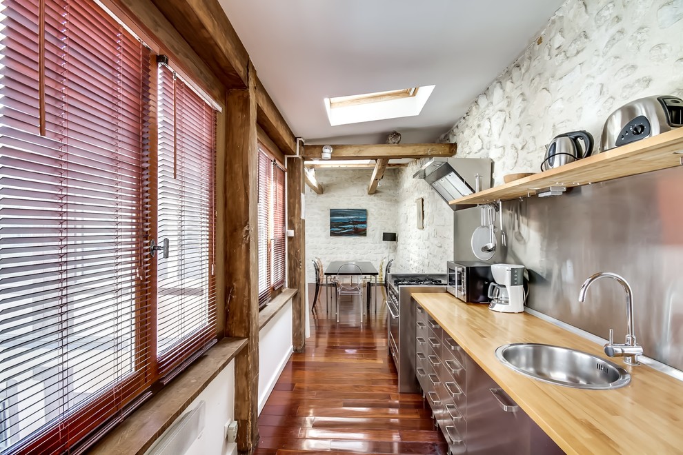 Bild på ett stort lantligt linjärt kök och matrum, med en enkel diskho, släta luckor, skåp i rostfritt stål, träbänkskiva, stänkskydd med metallisk yta, rostfria vitvaror och mellanmörkt trägolv