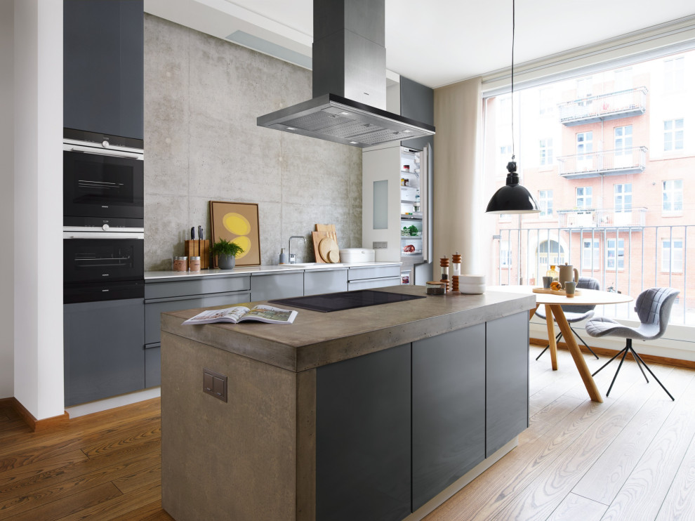 Cette image montre une cuisine ouverte parallèle et encastrable design avec des portes de placard grises, un plan de travail en béton, une crédence grise, un sol en bois brun, îlot, un sol marron et un plan de travail gris.