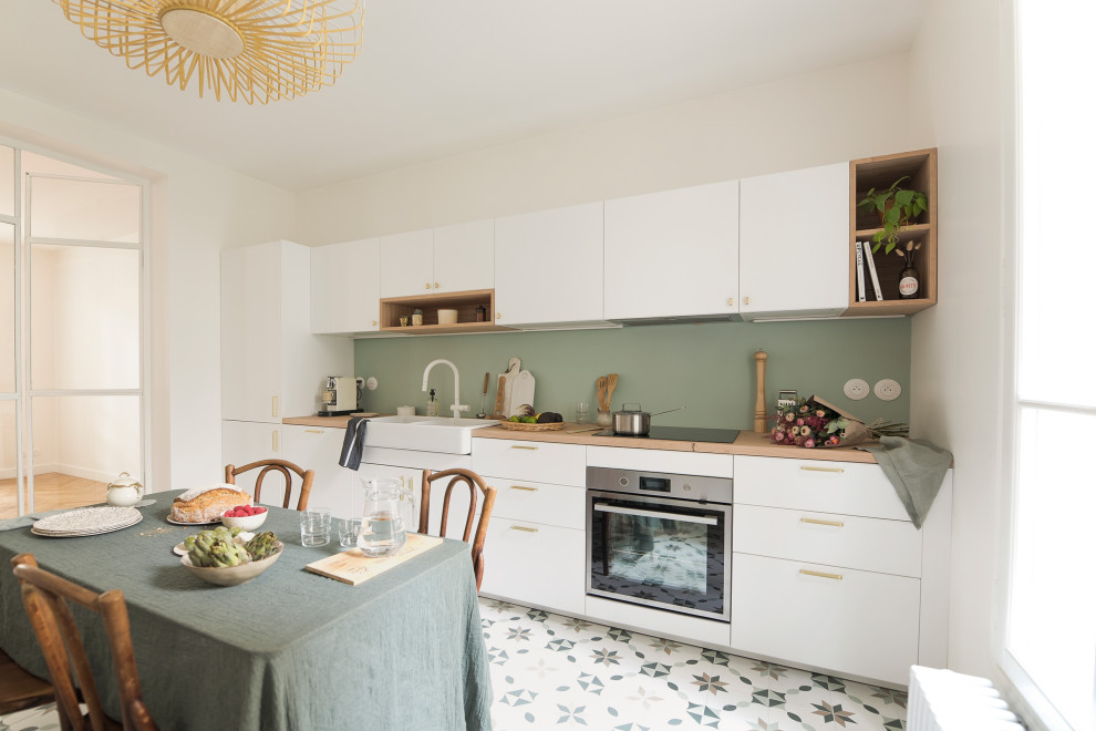 Foto på ett stort linjärt kök med öppen planlösning, med en dubbel diskho, släta luckor, vita skåp, träbänkskiva, grönt stänkskydd, cementgolv och flerfärgat golv