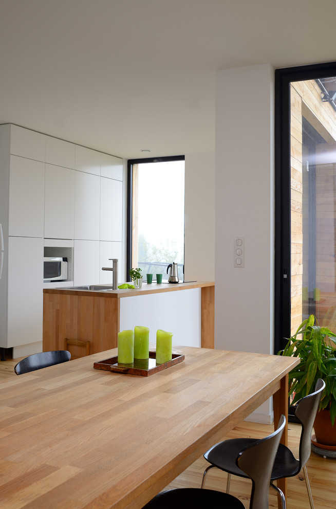 Свежая идея для дизайна: прямая кухня среднего размера, в белых тонах с отделкой деревом в современном стиле с обеденным столом, белыми фасадами и островом - отличное фото интерьера