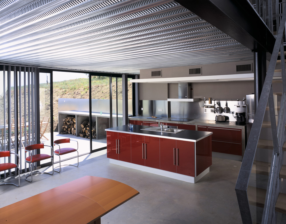 Inredning av ett industriellt mellanstort kök, med släta luckor, röda skåp, bänkskiva i betong, stänkskydd med metallisk yta, rostfria vitvaror, betonggolv och en köksö