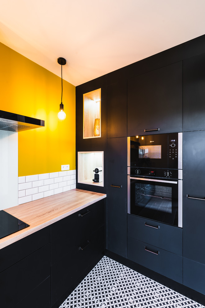 На фото: угловая кухня-гостиная среднего размера в стиле лофт с врезной мойкой, черными фасадами, деревянной столешницей, белым фартуком, фартуком из плитки кабанчик и черной техникой без острова