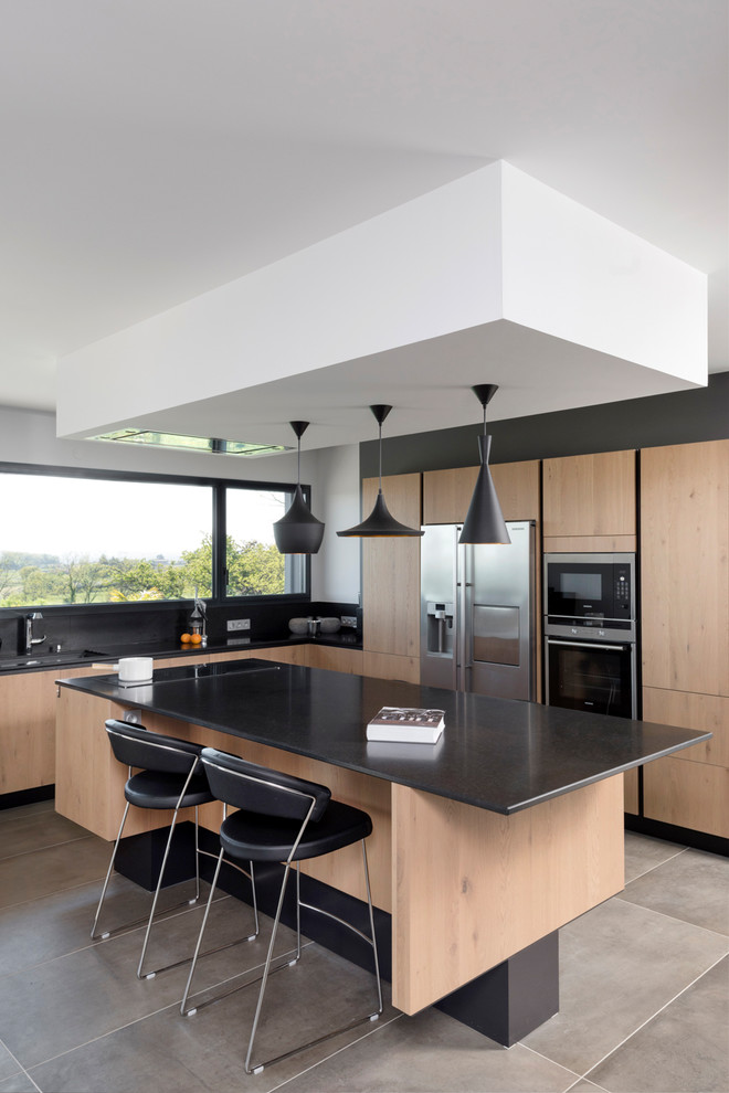 Moderne Küche in L-Form mit flächenbündigen Schrankfronten, hellen Holzschränken, Küchengeräten aus Edelstahl, Kücheninsel und grauem Boden in Lyon