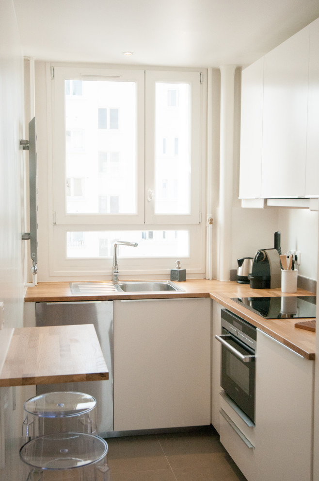This is an example of a small contemporary l-shaped kitchen in Paris with a built-in sink, flat-panel cabinets, white cabinets, wood worktops, black appliances, no island and grey floors.