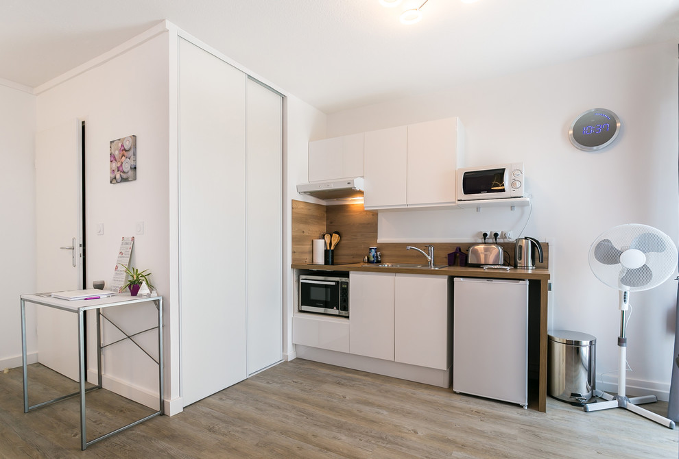 Photo of a contemporary single-wall open plan kitchen in Bordeaux with a built-in sink, flat-panel cabinets, white cabinets, wood worktops, brown splashback, wood splashback, white appliances, light hardwood flooring, no island and beige floors.