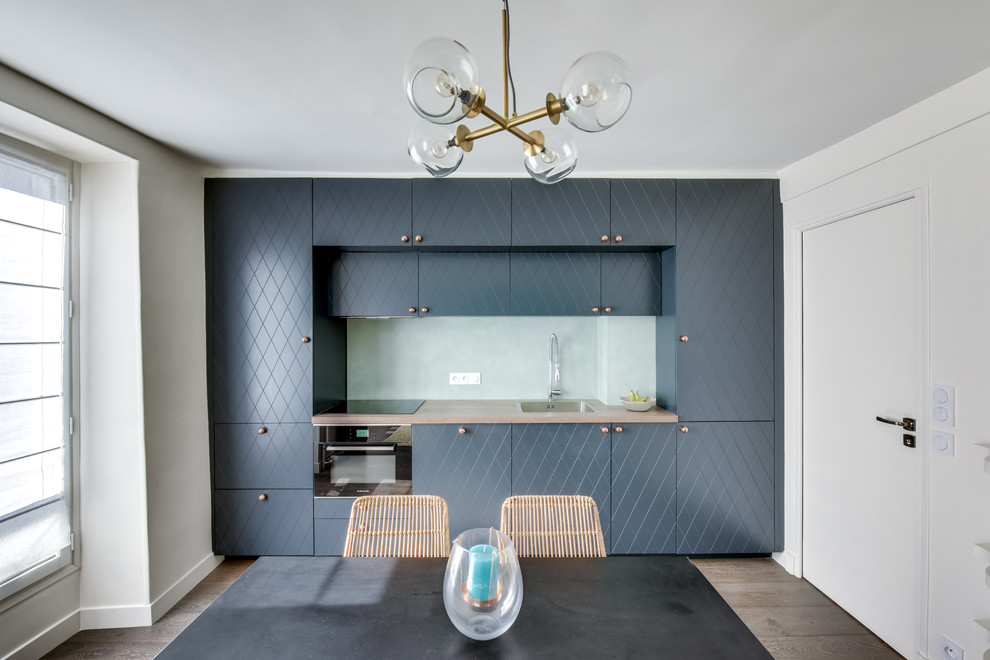 Einzeilige, Kleine Moderne Wohnküche ohne Insel mit Waschbecken, blauen Schränken, Glasrückwand, schwarzen Elektrogeräten, Arbeitsplatte aus Holz, Küchenrückwand in Blau und dunklem Holzboden in Paris