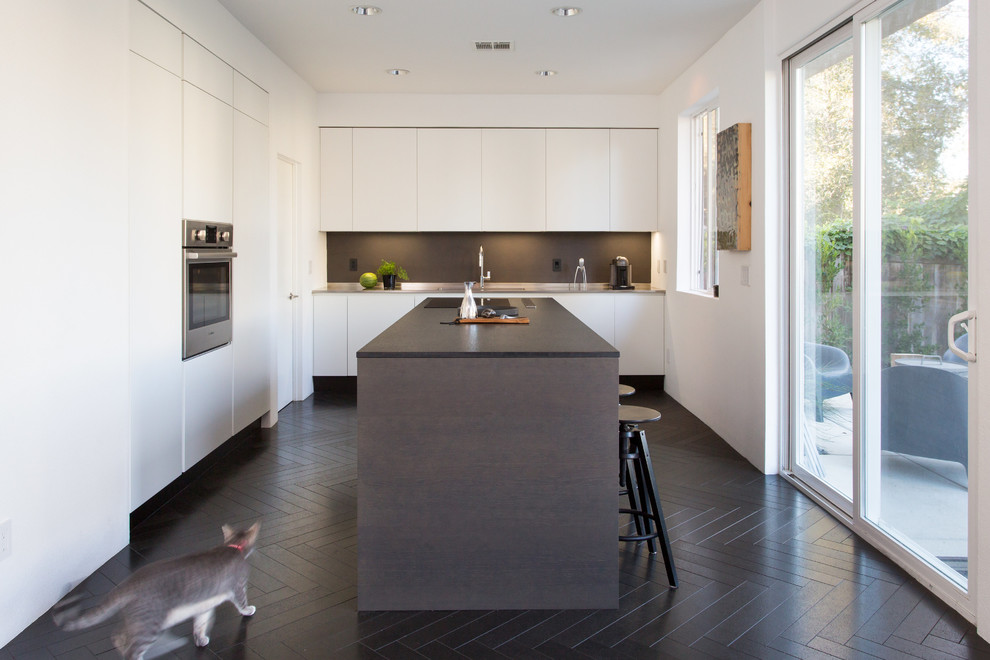 Modern inredning av ett svart svart l-kök, med släta luckor, vita skåp, svart stänkskydd, rostfria vitvaror, en köksö och svart golv