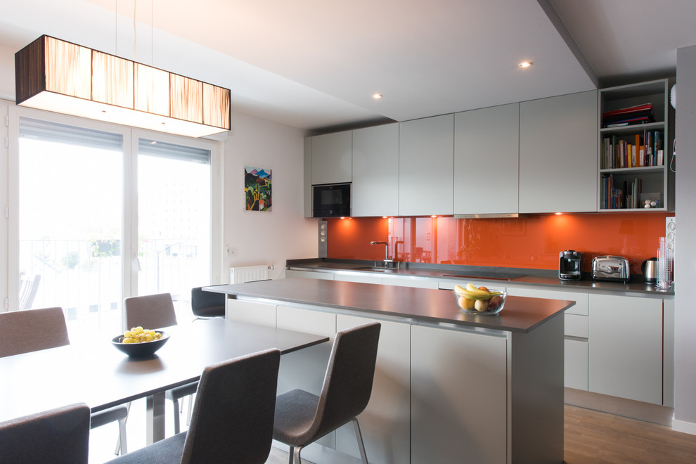 Idéer för att renovera ett stort funkis linjärt kök och matrum, med en integrerad diskho, luckor med profilerade fronter, grå skåp, bänkskiva i kvartsit, orange stänkskydd, glaspanel som stänkskydd, integrerade vitvaror, ljust trägolv och en köksö