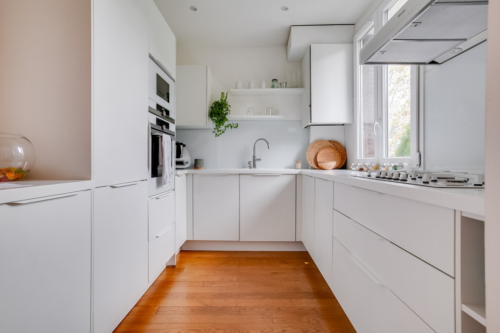 Inspiration for a small contemporary u-shaped open plan kitchen in Paris with white cabinets, quartz worktops, white splashback, glass tiled splashback, white appliances, bamboo flooring, brown floors, white worktops, a built-in sink, flat-panel cabinets and no island.