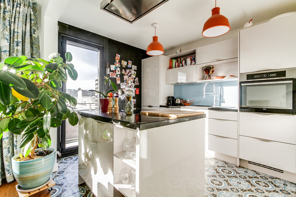 Cette image montre une cuisine nordique avec un placard à porte plane, une crédence bleue, une crédence en feuille de verre, îlot, un sol multicolore et plan de travail noir.