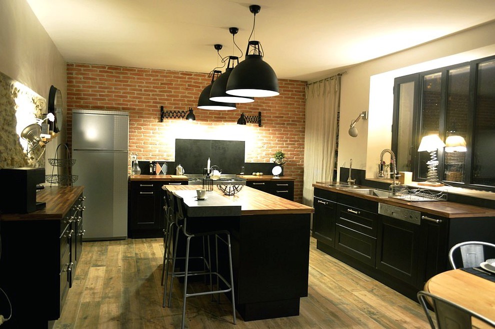 Inredning av ett industriellt mellanstort beige beige kök, med en dubbel diskho, luckor med infälld panel, svarta skåp, träbänkskiva, svart stänkskydd, stänkskydd i keramik, rostfria vitvaror, laminatgolv, en köksö och beiget golv