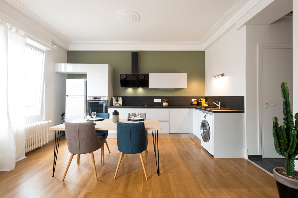 Medium sized scandi l-shaped open plan kitchen in Grenoble with a single-bowl sink, flat-panel cabinets, white cabinets, granite worktops, black splashback, light hardwood flooring, no island, black worktops, white appliances and brown floors.